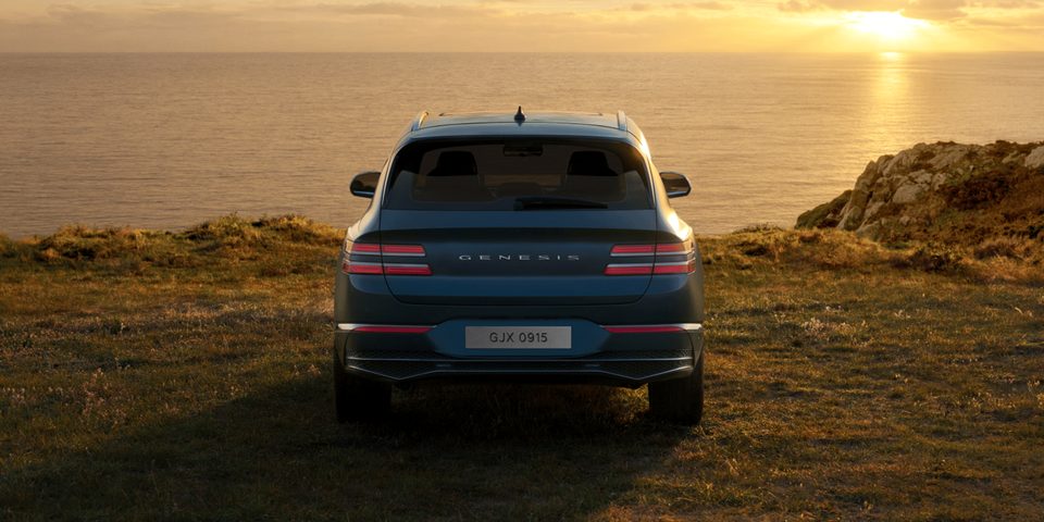 Rear view of regular GV80 facing a sunset coast; rear lamps and GENESIS lettering visible.